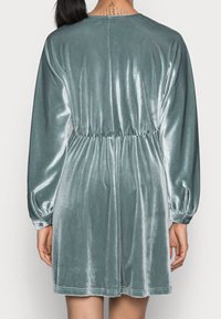 Sametové šaty v tlumené barvě teal s dlouhými rukávy, elastickým pasem a mírně rozšířenou sukní. Hladká textura a minimální detaily.