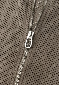 Sølv lynlås delvist åbnet på taupe perforeret stof med små firkantede huller, der viser tekstur og detaljer af lynlåsens tænder.