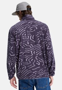 Fleece pullover in dark purple with abstract white wave patterns. Collared design, long sleeves, relaxed fit, and soft texture.