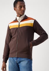 Braune Zip-Jacke mit orangefarbenen, weißen und gelben horizontalen Streifen über der Brust. Mit hohem Kragen und seitlichen Taschen.