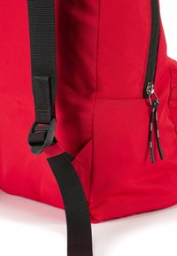 Zaino di tessuto rosso con spallacci neri, una tasca con zip e cuciture evidenti. Materiale testurizzato con dettagli hardware a contrasto.