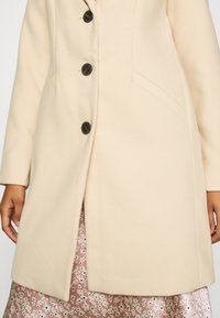 Cappotto in lana beige con taglio dritto, cinque bottoni scuri e una tasca laterale. Presenta una texture liscia e viene indossato sopra una gonna floreale.