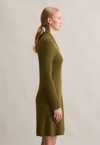 Abito a coste verde oliva con maniche lunghe, colletto e orlo svasato, caratterizzato da dettagli di bottoni discreti lungo il décolleté.