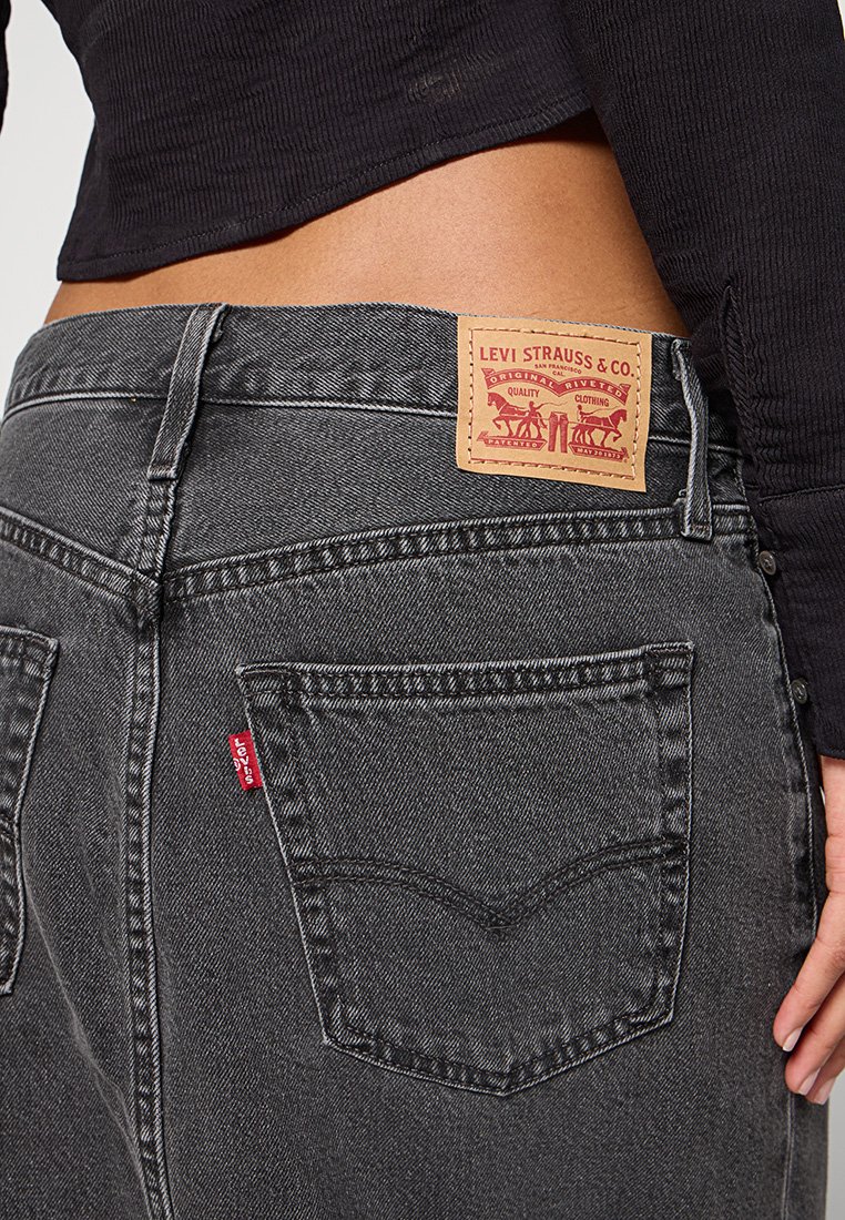 Jeans de mezclilla negra con un lavado oscuro, bolsillos traseros cosidos y una etiqueta de cuero marrón con texto y gráficos rojos.