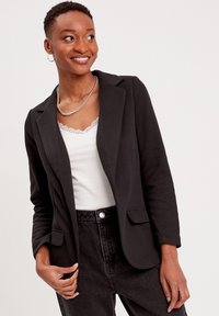 Blazer noir ajusté avec revers crantés, fermeture à un bouton, deux poches avant et un tissu lisse et léger. Associé à un top blanc avec des dentelles.