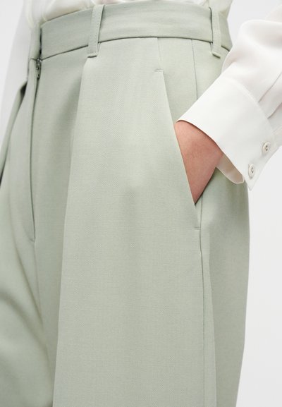 Ανοιχτό πράσινο παντελόνι με υφή υφάσματος, που διαθέτει πιέτες και πλαϊνές τσέπες. Ένα λευκό πουκάμισο με μακριά μανίκια και μανσέτες με κουμπιά φαίνεται μερικώς.