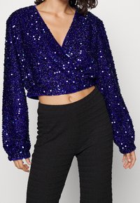 Top corto in paillettes viola con maniche lunghe, design a scollo a V e tessuto testurizzato, abbinato a pantaloni neri a vita alta e arricciati.