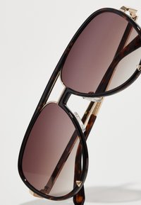 Gafas de sol con una montura de acetato de carey oscuro, acentos metálicos dorados y lentes marrones degradados. Presentan una forma geométrica audaz.