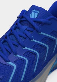 Primer plano de una zapatilla azul K-Swiss con malla texturizada, cordones azules a juego y superposiciones en forma de onda de color azul claro en el lateral.