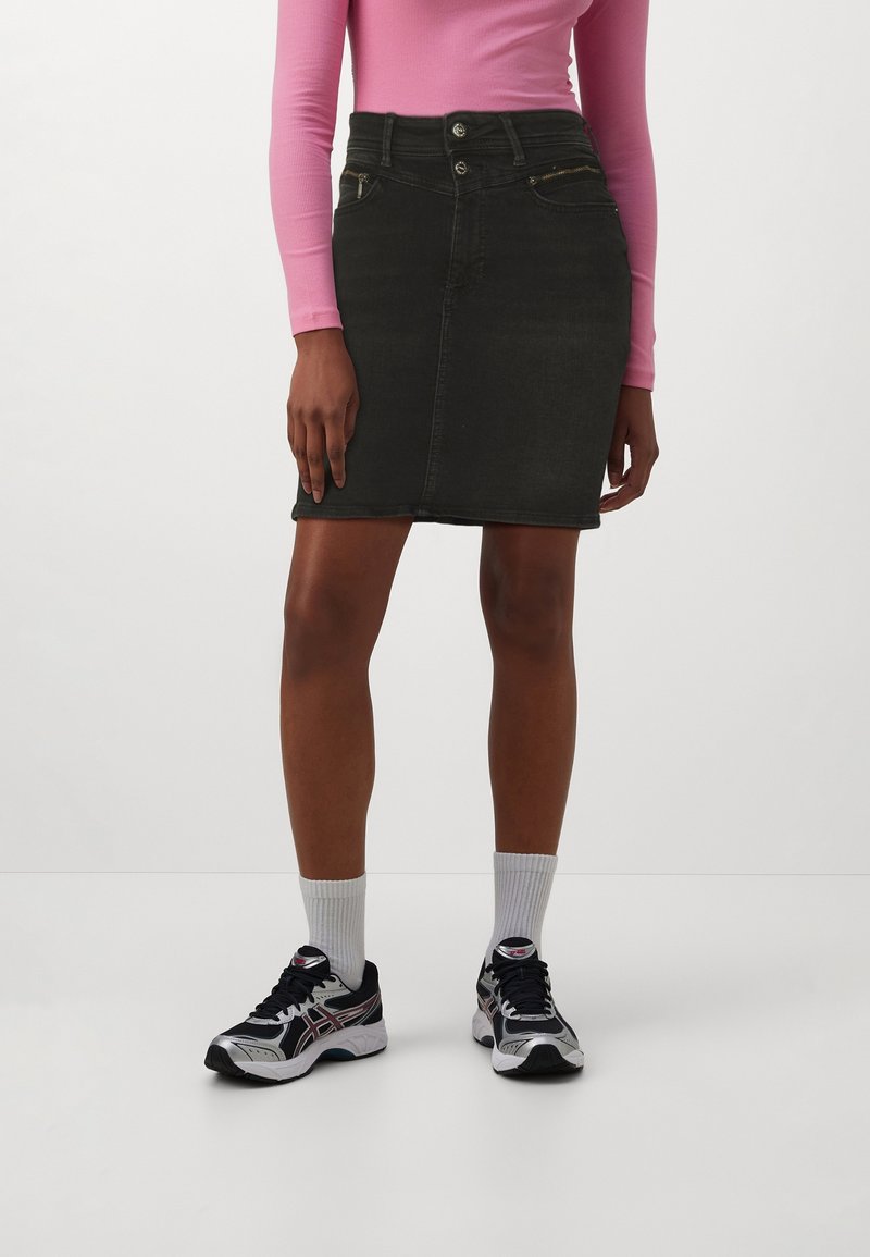 Femme portant une jupe en jean noire, un haut à manches longues rose, des chaussettes blanches et des chaussures de course noires et blanches, debout devant un fond uni.