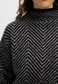 Schwarz-cream gestreifter Pullover mit Fischgrätmuster, strukturiertem Strick und hohem Kragen. Nahaufnahme des oberen Kleidungsstücks.