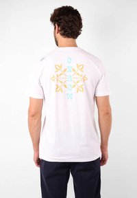 T-shirt blanc en coton avec un design graphique de motifs de feuilles bleues et jaunes au dos. Manches courtes et encolure ronde.