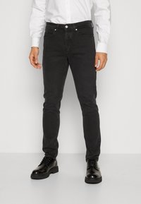 Hombre con jeans ajustados negros, zapatos de cuero negros con cordones y una camisa blanca de botones, de pie contra un fondo liso.