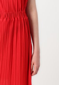Vestido plissado vermelho com cintura elástica, apresentando pregas verticais e uma textura de tecido suave. O tecido cai suavemente.