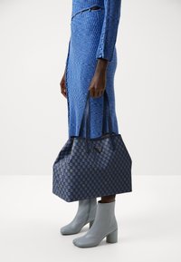 Osoba ubrana w niebieską sukienkę w prążki i szare botki do kostek trzyma dużą niebieską torbę w wzory z podwójnymi uchwytami.