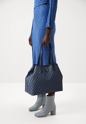 Henkilö, joka käyttää sinistä ribbitettyä mekkoa ja harmaita nilkkureita, pitää käsissään suurehkoa sinistä kuviolaukku kaksinkertaisilla kahvoilla.