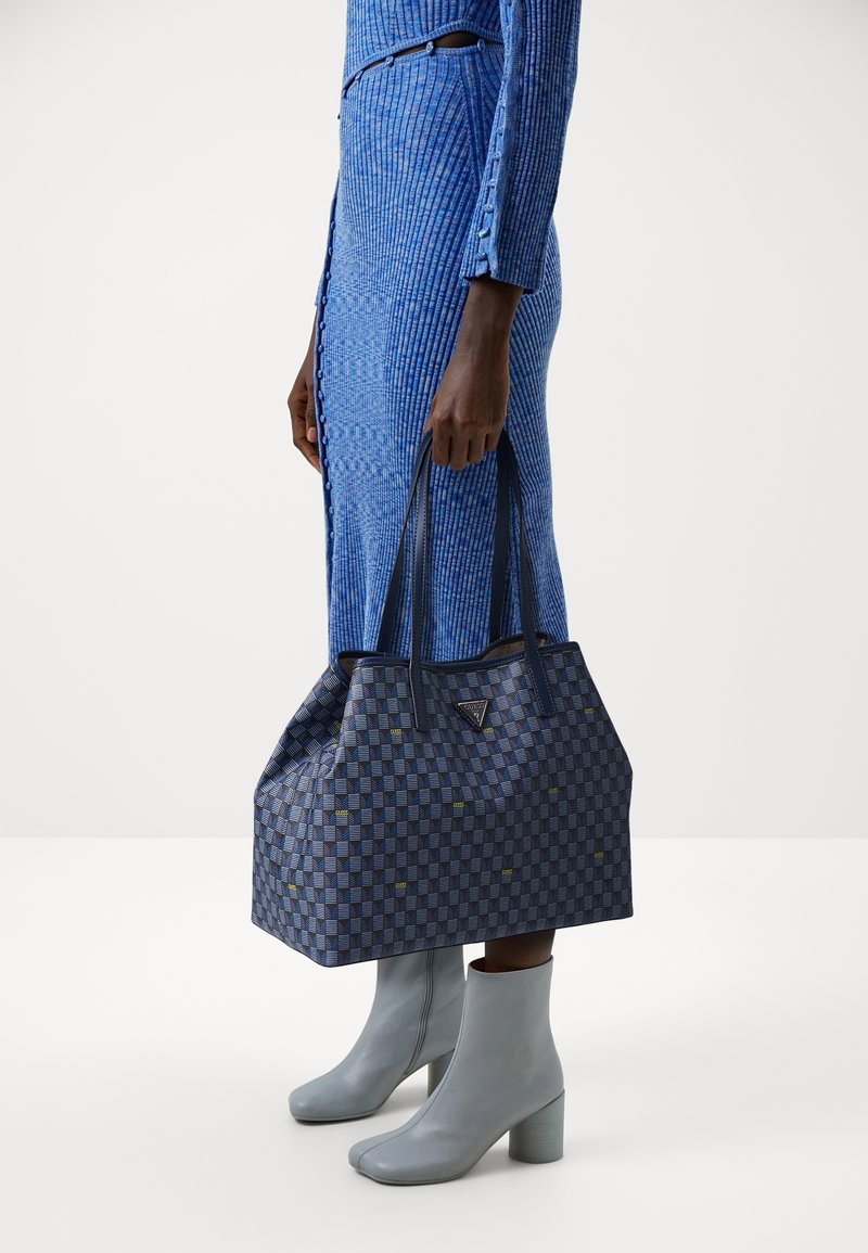 Osoba ubrana w niebieską sukienkę w prążki i szare botki do kostek trzyma dużą niebieską torbę w wzory z podwójnymi uchwytami.