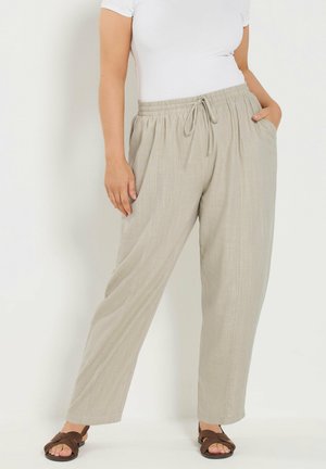 Frau trägt beige, locker sitzende Leinenhose mit elastischem Kordelzug in der Taille, weißes figurbetontes Kurzarmoberteil und braune Sandalen, steht mit der Hand in der Tasche.