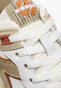 Zapatilla blanca que combina materiales de cuero y malla, con acentos en beige y patrones gráficos en naranja en la lengüeta. Cordones blancos.