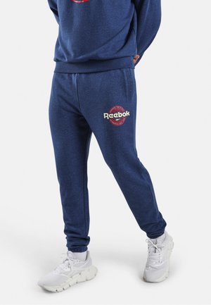 Persoon die marineblauwe Reebok joggingbroek en bijpassende trui draagt, staat met de handen achter de rug, witte sportschoenen zichtbaar.