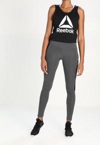 Top in tessuto nero di Reebok con logo bianco, abbinato a leggings grigi aderenti. Scarpe da ginnastica nere con suole testurizzate completano l'outfit.
