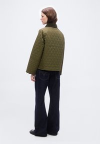 Γυναίκα που φοράει σκούρο μπλε τζιν παντελόνι φαρδιάς γραμμής και πράσινο καπιτονέ μπουφάν με καφέ γιακά, κοιτάζοντας πλαγίως σε λευκό φόντο.