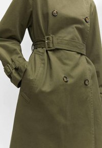 Płaszcz trencz w oliwkowej zieleni z dwurzędowym zapięciem na guziki, pasem w tym samym kolorze i kieszeniami po bokach. Wykonany z lekkiego materiału o gładkiej fakturze.