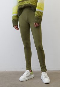 Olivgröna leggings med en figurnära design, slät textur och sidodragkedja, parat med vita sneakers med gröna detaljer.