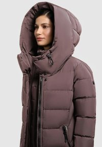 Jakna od nabranog materijala u mauve boji s velikim, podstavljenim kapuljačom. Ima prednji patentni zatvarač, bočne džepove s patentnim zatvaračem i detalje na dugmad. Glatka tekstura.