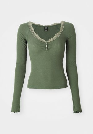Olivgrünes, geripptes Langarmoberteil mit V-Ausschnitt, Spitzeneinsatz, Knopfleiste mit drei Knöpfen und Rüschensaum. Weicher Stoff mit figurbetontem Schnitt.
