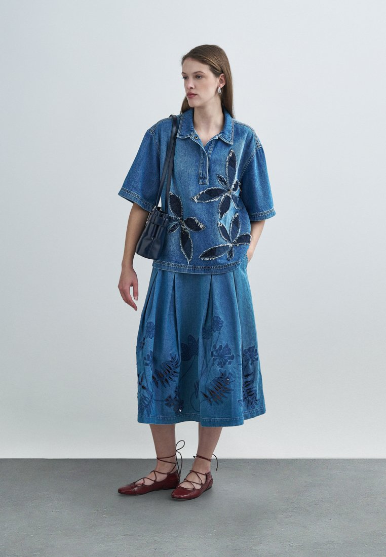 Tenue en denim comprenant une chemise à manches courtes avec broderies florales et une jupe plissée aux motifs similaires. Ballerines marron et petit sac.