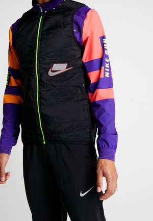 Mand iført sort Nike vest med neon grøn lynlås over en lilla og orange langærmet trøje og sorte Nike bukser.