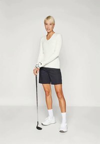 Γυναίκα με κοντά ξανθά μαλλιά, φορώντας λευκή μπλούζα με μακριά μανίκια, μαύρες βερμούδες, λευκές κάλτσες και αθλητικά παπούτσια, κρατώντας μια ράμπα γκολφ και στέκεται σε απλό φόντο.