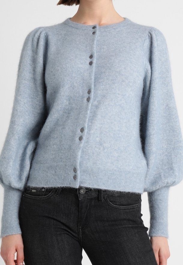 Femme portant un cardigan duveteux bleu clair avec manches bouffantes et devant à boutons noirs, associé à un jean noir.