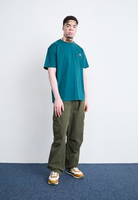 Green cotton t-shirt with logo, paired with olive cargo pants. Footwear features orange and white sneakers with green accents.