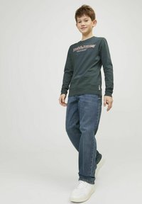 Tamnozeleni dukserica dugih rukava s logotipom "Jack & Jones", u kombinaciji s plavim trapericama i bijelim tenisicama u stojećem položaju. Glatka tekstura tkanine.