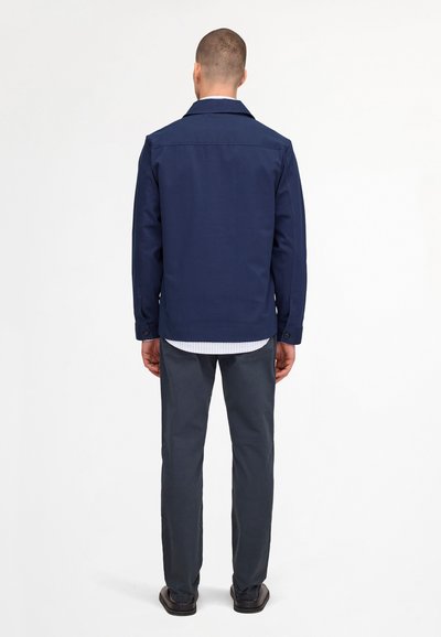 Uomo in piedi di spalle, indossa una giacca blu navy, una camicia a righe, pantaloni scuri e scarpe nere su uno sfondo bianco uniforme.