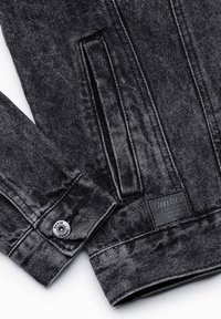 Manche de veste en denim noir et poche latérale avec un bouton métallique portant la mention « DENIM OMBRE » et une étiquette de marque noire cousue près de l’ourlet.