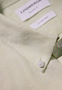 Lniania koszula w jasnym zielonym kolorze z wyraźnym kołnierzykiem, z pojedynczym białym guzikiem i metkami pielęgnacyjnymi wszytymi w materiał.