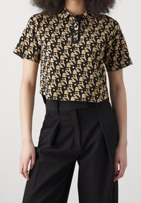 Femme portant un polo à manches courtes noir et beige à motifs, rentré dans un pantalon noir plissé taille haute, debout devant un fond blanc.