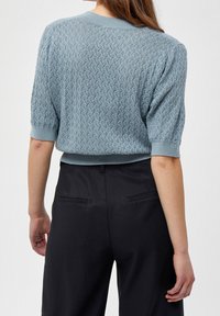Helles blauer Strickpullover mit kurzen Ärmeln, der ein strukturiertes Rautenmuster und einen gerippten Saum aufweist. Dunkle Hosen ergänzen das Outfit.