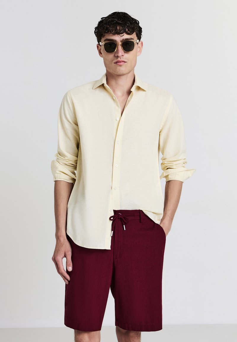 Chemise à boutons jaune clair en lin, associée à un short bordeaux à cordon. Des lunettes de soleil rondes complètent le look.