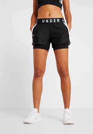 Kobieta ubrana w czarne, warstwowe szorty Under Armour oraz białe trampki stoi z rękami w kieszeniach na jednolitym białym tle.