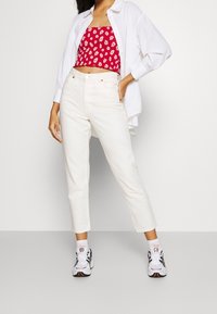 Žena v bílých teniskách, bílých džínách s vysokým pasem, červeném květinovém topu a volné bílé košili stojí proti jednoduchému pozadí.