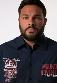 Camicia blu navy con bottoni, caratterizzata da grafiche ricamate, tra cui "ESPLORA IL MONDO" e "AVVENTURA MONTANA", con cuciture dettagliate.