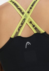 Reggiseno sportivo nero con design a incrocio sulla schiena. Presenta spalline in rete gialle con un motivo. Include un dettaglio con il logo nella parte bassa della schiena.