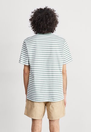 Person med krøllet mørkt hår iført en hvid og grønstribet kortærmet skjorte og beige shorts, vist bagfra.