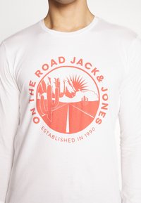 Valkoinen pitkähihainen paita, jossa on punainen graafinen kuvio, joka esittää tietä, kaktuksia, vuoria ja tekstit "JACK & JONES" sekä "PERUSTETTU 1990".