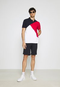 Polo con un design a blocchi di colore navy, rosso e bianco, abbinato a pantaloni corti scuri. Modello che indossa occhiali da sole e sneaker bianche.