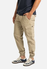 Hombre vistiendo pantalones de chándal cargo beige y zapatillas azul marino con rayas blancas, de pie con una mano en el bolsillo contra un fondo blanco.